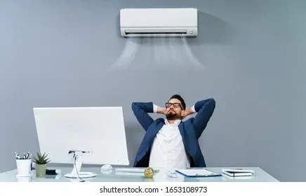A man in a suit relaxing at his desk beneath an air conditioning unit.